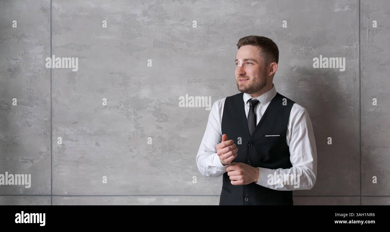 homme en vêtements d'affaires se tient contre le mur, ajustant les poignets de sa chemise, avec un look concentré. L'arrière-plan neutre et l'éclairage doux créent un atm Banque D'Images