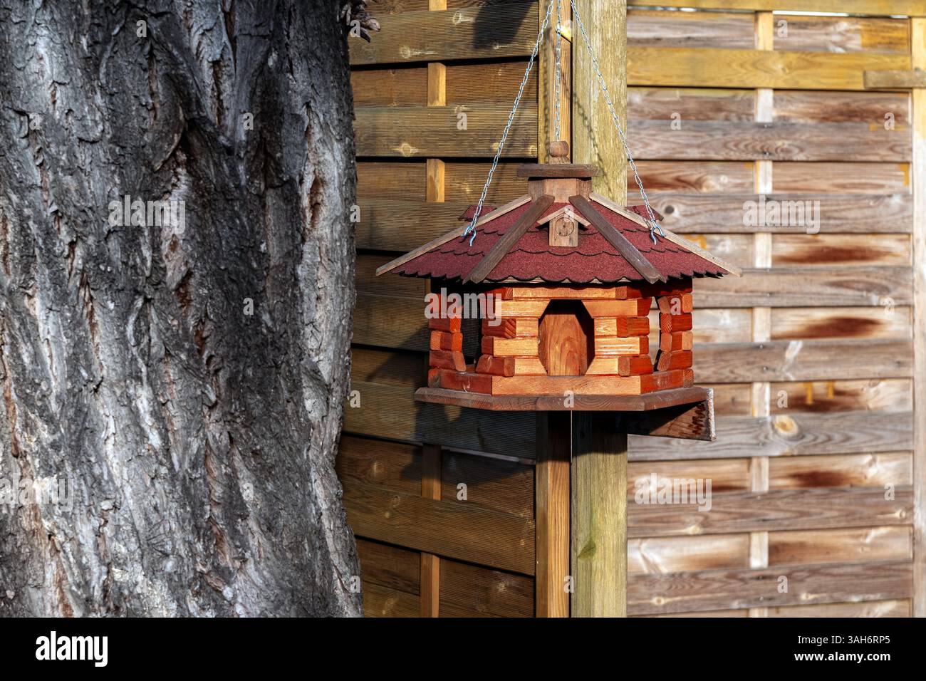 Mangeoire à oiseaux en bois en bois sous la forme d'une maison suspendue à une clôture à côté d'un arbre. Banque D'Images