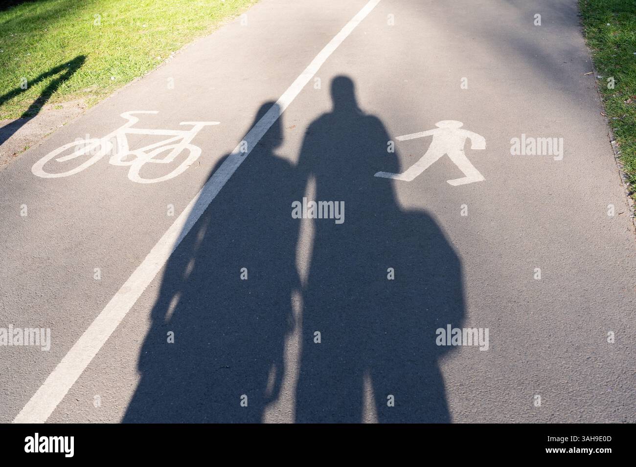Un couple marchant côte à côte projetant de longues ombres dans le soleil de fin d'après-midi sur un sentier / piste cyclable séparé à Eastrop Park, Basingstoke, Royaume-Uni Banque D'Images