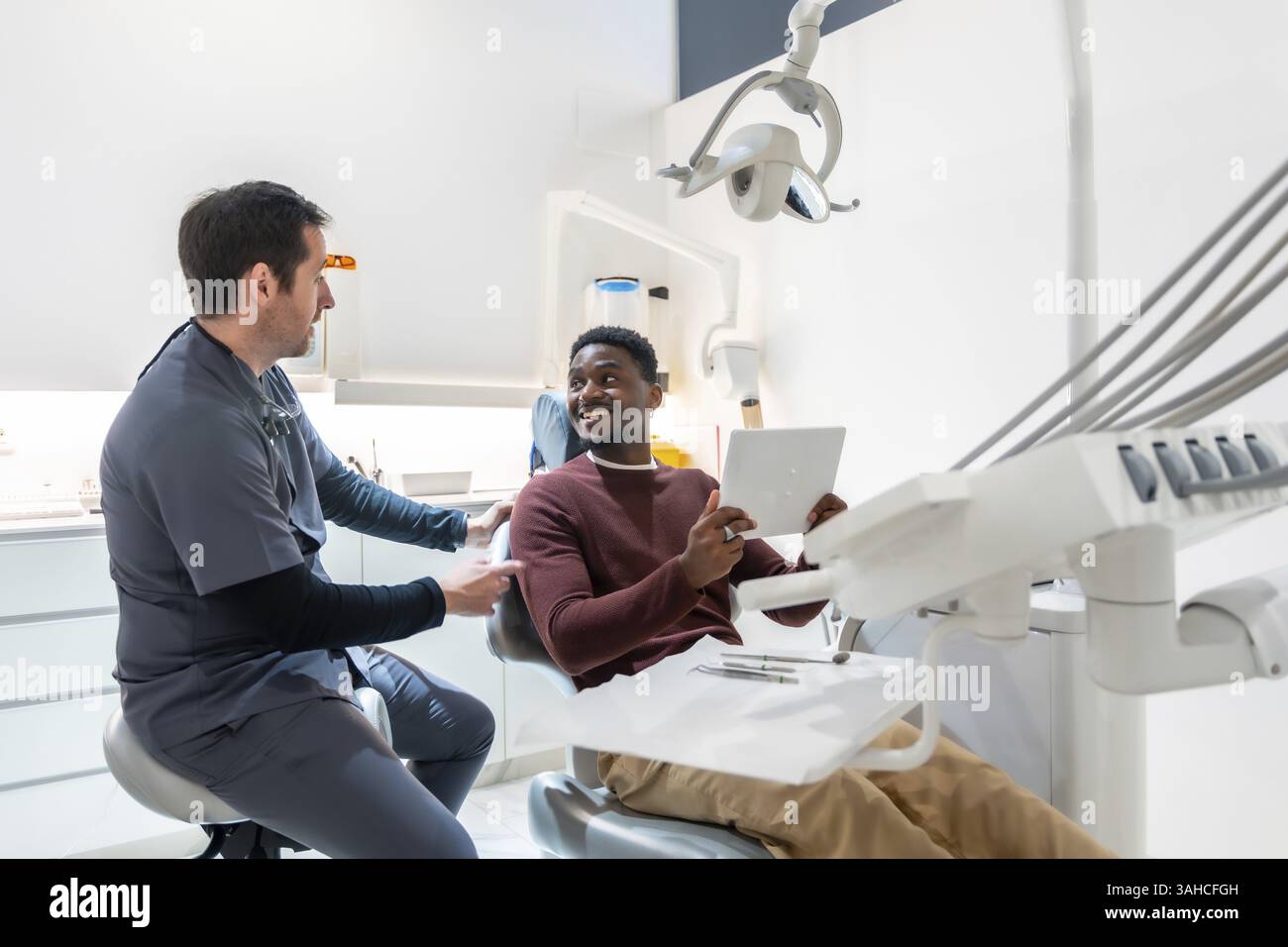 Dentiste montre les résultats d'un examen dentaire à un patient souriant sur une tablette dans une clinique dentaire moderne et lumineuse Banque D'Images