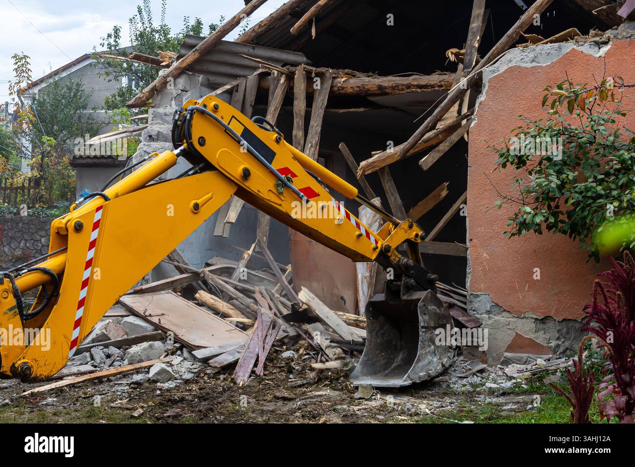 Une pelle jaune démolit activement un bâtiment endommagé avec son godet. Des débris et des structures brisées entourent le site. La poussière remplit l'air comme t Banque D'Images