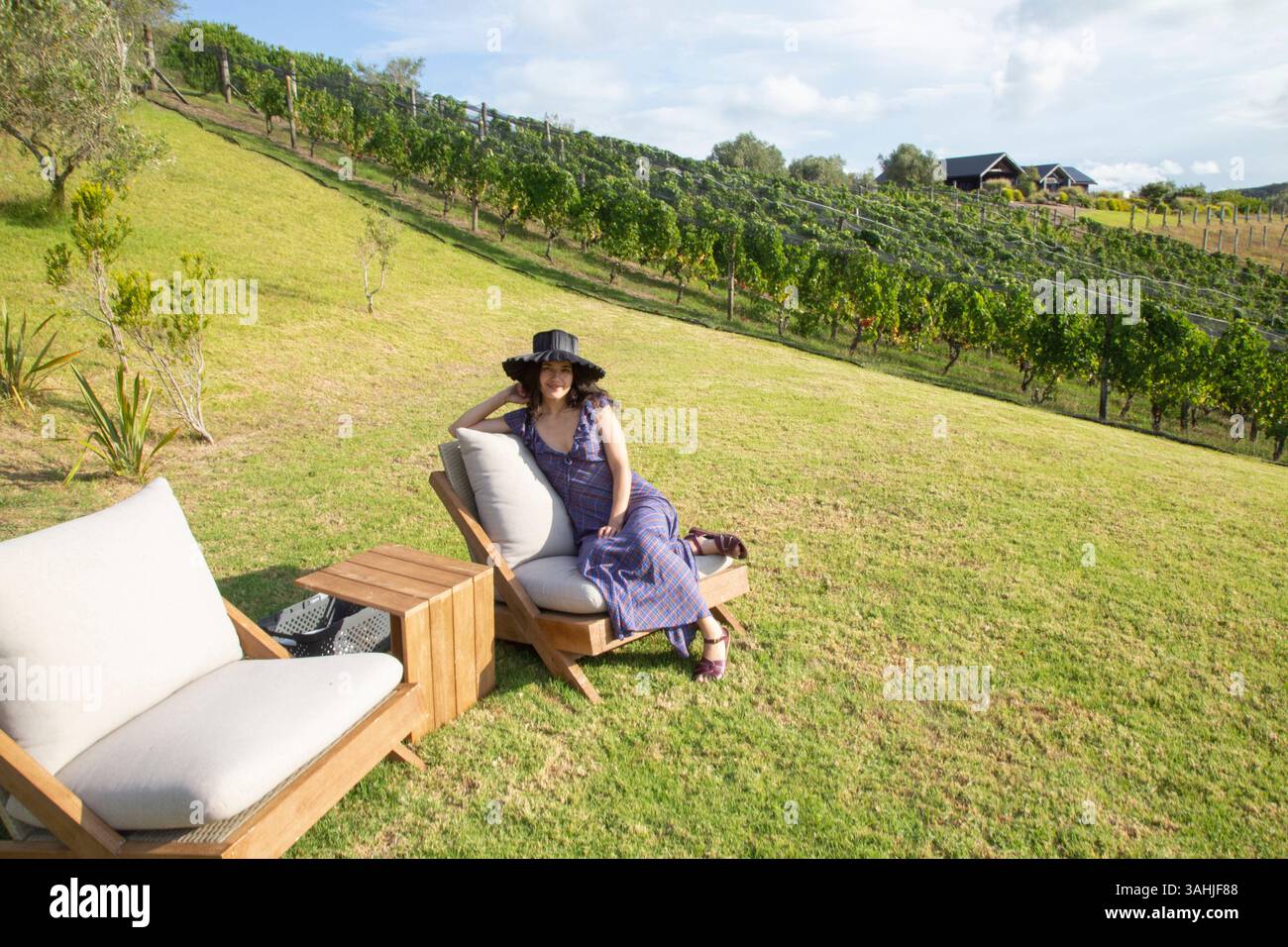 Femme dans une robe rayée et chapeau se détend sur une chaise longue dans un paysage viticole ensoleillé. Île de Waiheke, Auckland, Nouvelle-Zélande Banque D'Images