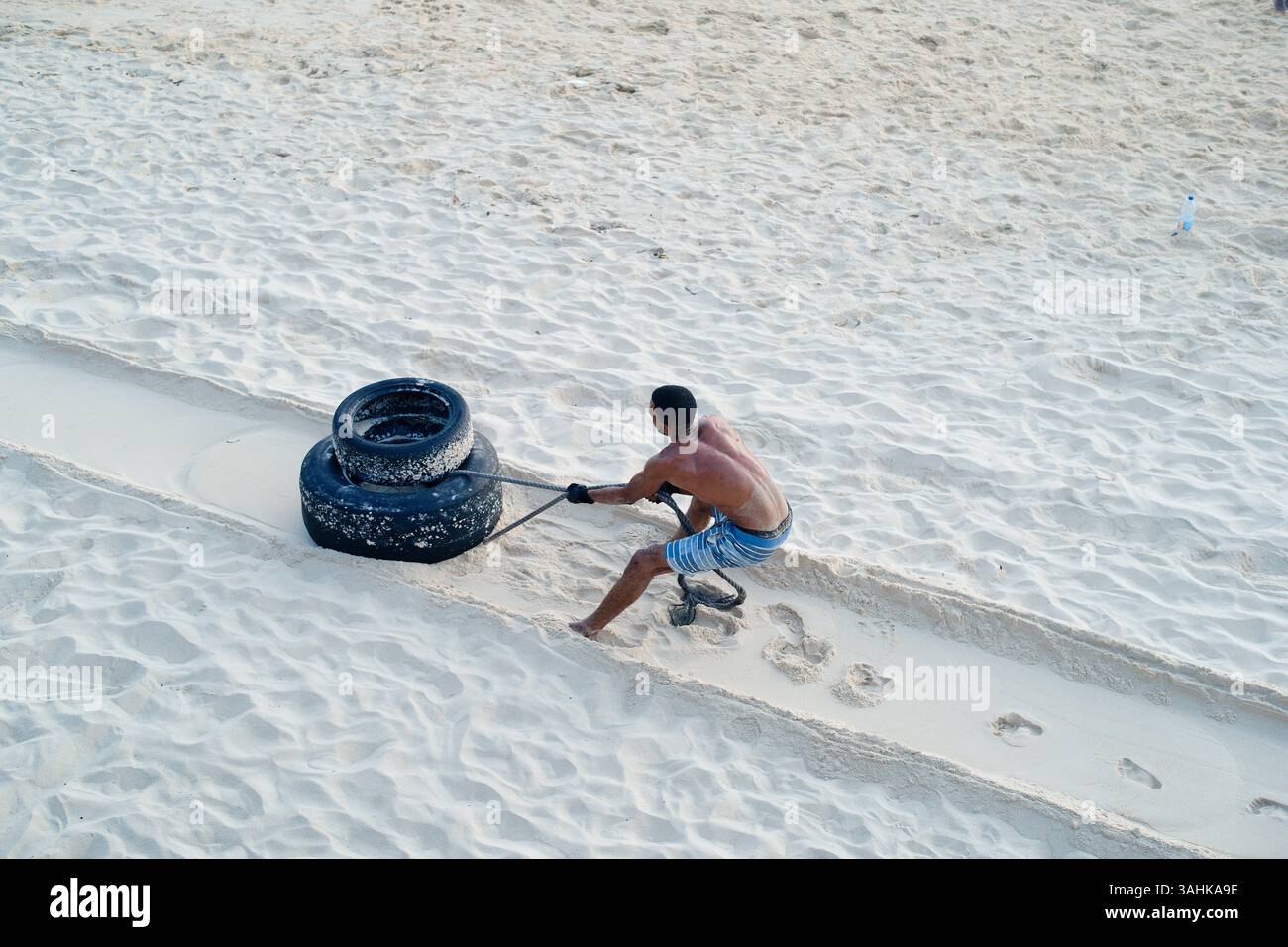 Un homme traîne deux gros pneus à travers le sable sur une plage ensoleillée, laissant une traînée derrière. Tanzanie, Afrique Banque D'Images