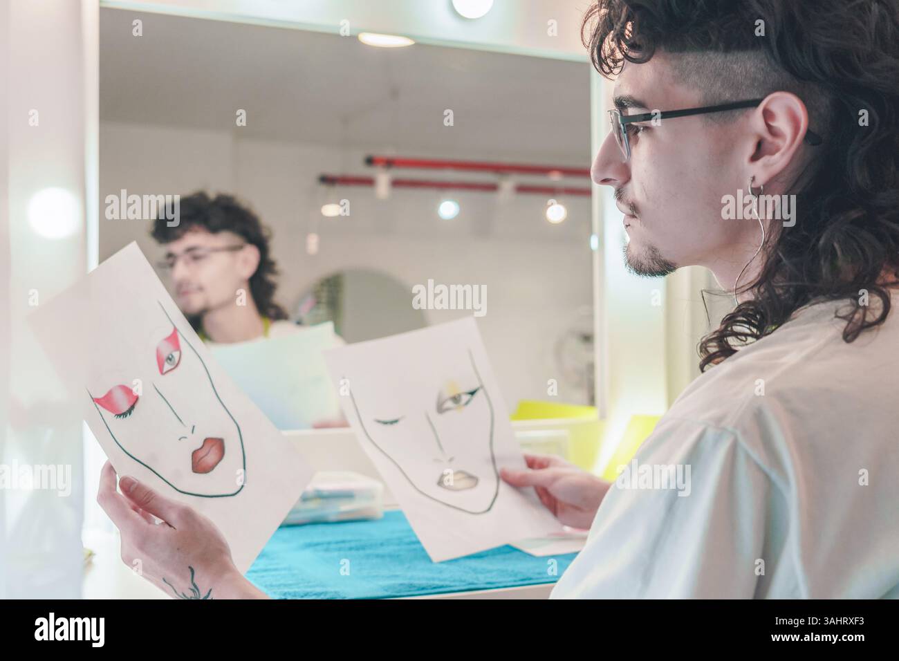 Jeune maquilleur masculin comparant différentes cartes faciales tout en étant assis devant un miroir dans un salon de beauté lumineux, mettant en valeur la diversité et Banque D'Images