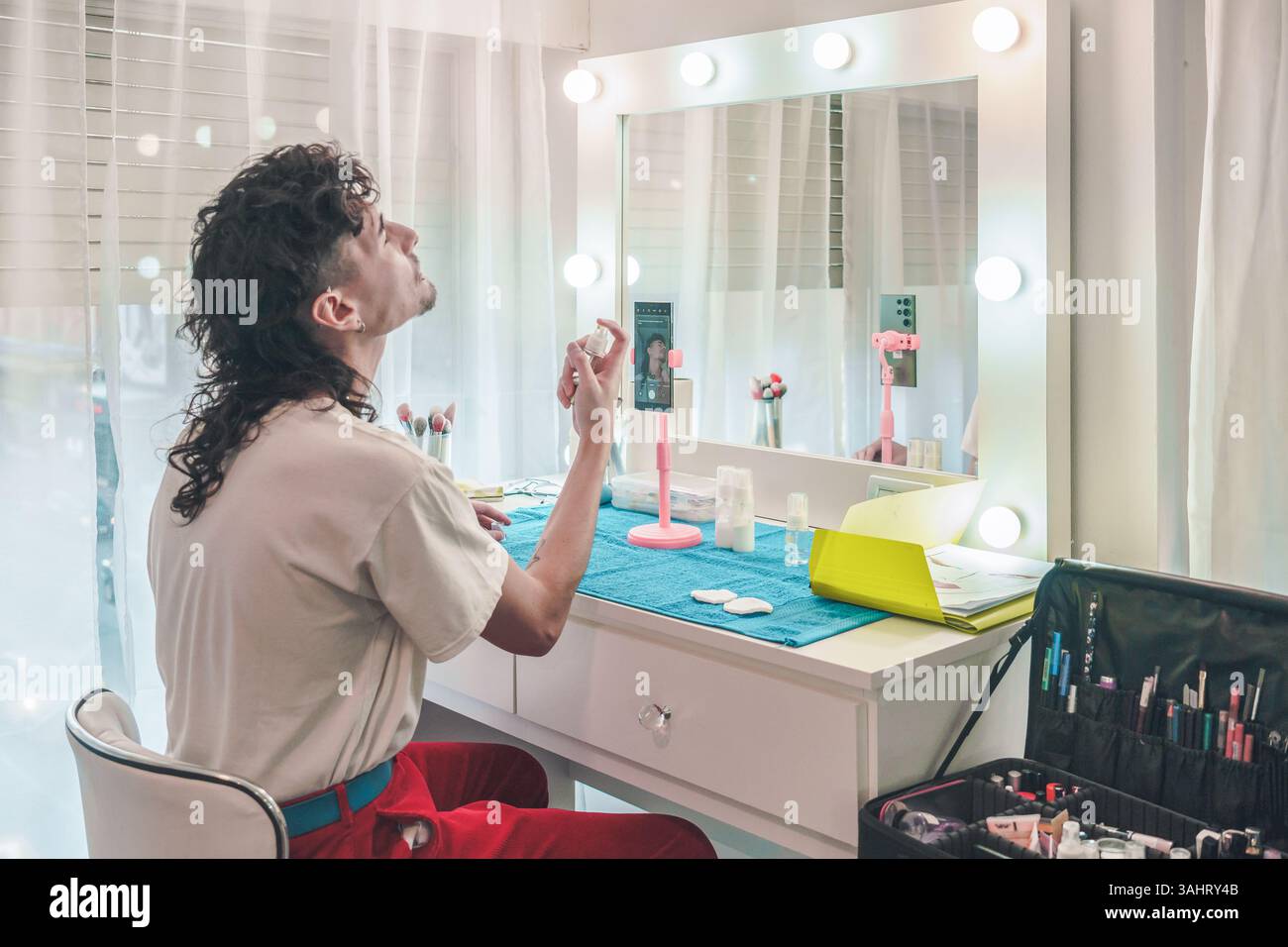 Jeune maquilleur masculin pulvérisant le spray de réglage sur son visage tout en enregistrant un tutoriel de maquillage avec son smartphone devant un miroir avec des lumières LED Banque D'Images