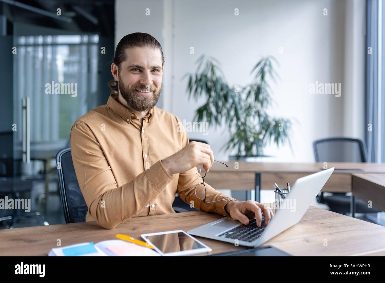 Un homme d'affaires souriant à son bureau, tenant des lunettes, travaille sur son ordinateur portable dans un cadre de bureau moderne. L'homme regarde directement la caméra, rayonnant de confiance. Banque D'Images