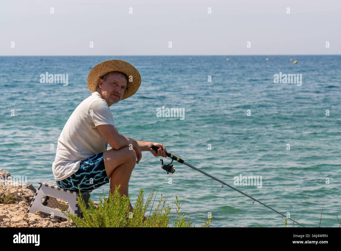 Homme pêcheur dans le chapeau est assis sur le bord de la mer avec la canne à pêche dans ses mains. Journée ensoleillée d'été. Vacances tranquilles à la mer, passe-temps. Banque D'Images