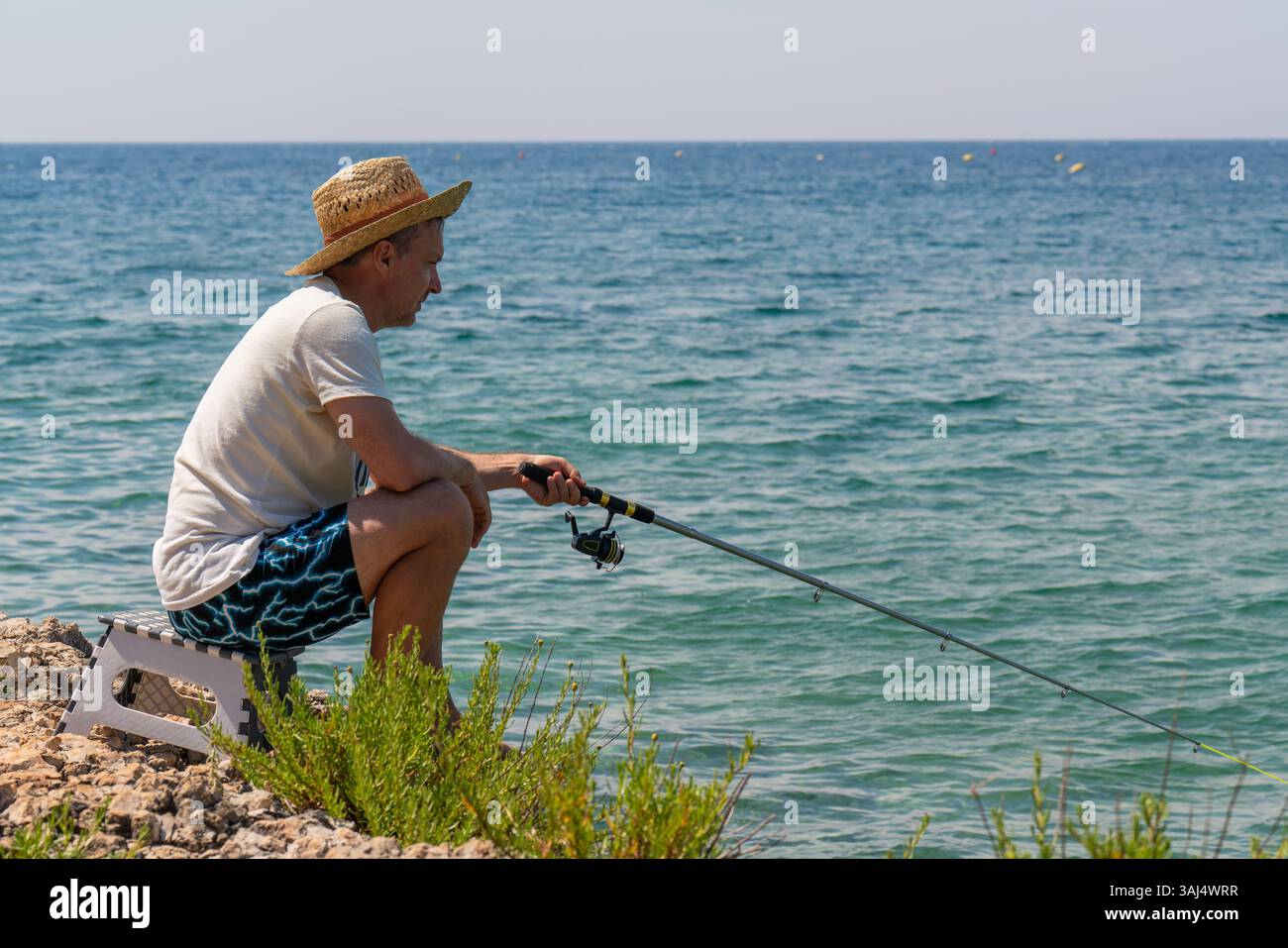 Homme pêcheur dans le chapeau est assis sur le bord de la mer avec la canne à pêche dans ses mains. Journée ensoleillée d'été. Vacances tranquilles à la mer, passe-temps. Banque D'Images
