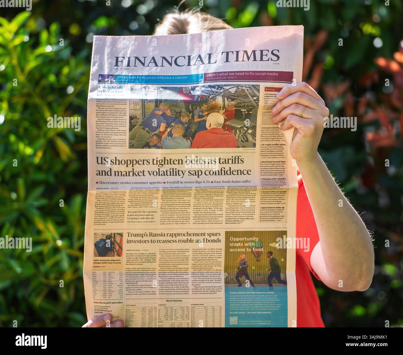 Paris, France - 9 avril 2025 : une femme en chemise rouge lit le Financial Times dans un jardin verdoyant, avec les gros titres sur les réductions des dépenses de consommation américaines, les effets tarifaires et les réactions des investisseurs aux relations avec la Russie Banque D'Images