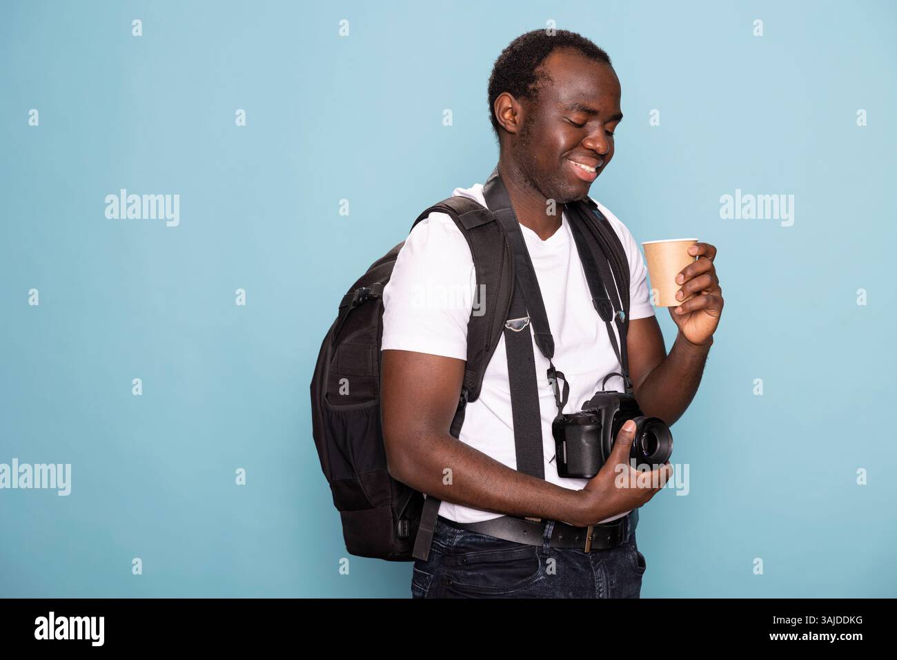 Homme afro-américain touche son objectif de caméra, souriant en regardant la tasse jetable dans sa main. Photographe masculin noir avec un sac à dos et un reflex numérique regarde son café avec une expression de contenu. Banque D'Images