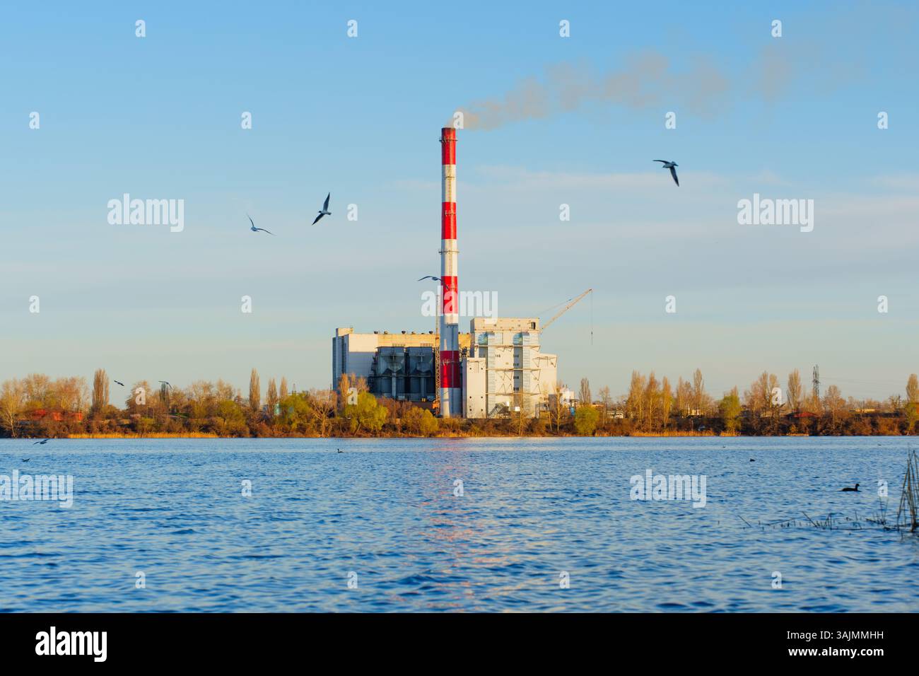 Usine moderne d'incinération des déchets située au bord d'un plan d'eau serein, mettant en évidence les émissions de fumée et l'environnement naturel, mettant l'accent sur la gestion des déchets a Banque D'Images
