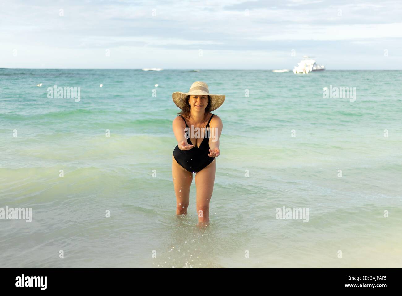 Une femme dans un maillot de bain noir se tient dans l'eau peu profonde, tendant la main avec les deux mains et souriant. La plage s'étend derrière elle comme le soleil se couche, créatine Banque D'Images