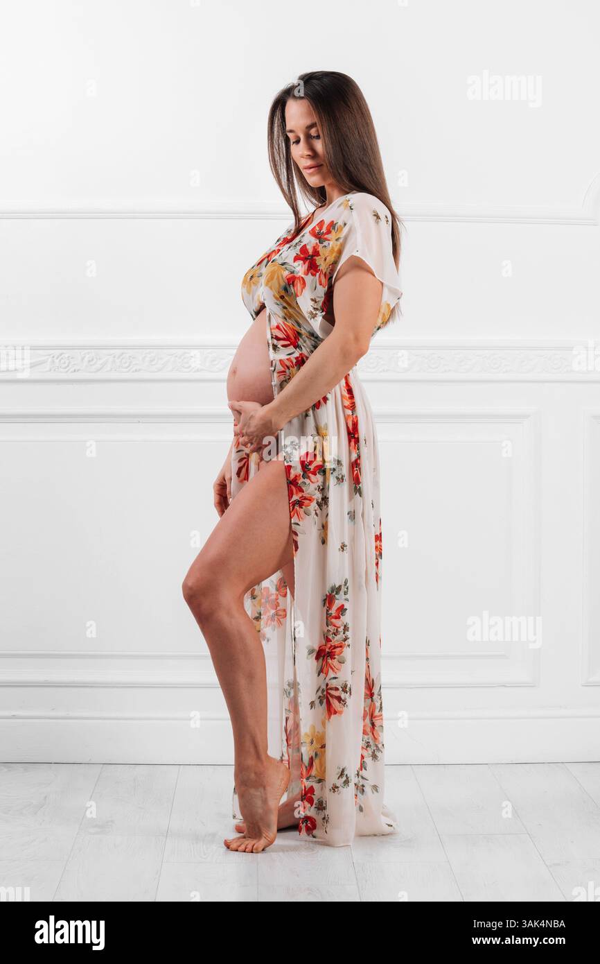 Mannequin enceinte portant une robe fleurie, posant dans un studio et touchant son ventre Banque D'Images