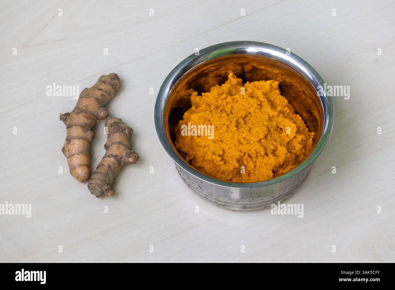 La pâte de curcuma cru dans un bol en acier repose sur une surface en bois clair, avec deux racines de curcuma cru entières placées à côté. Banque D'Images