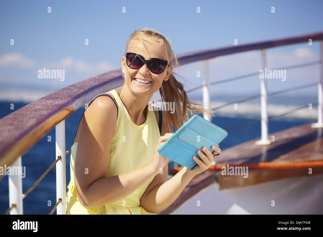 Jeune femme souriante dans des lunettes de soleil tenant un ordinateur tablette et regardant quelque part tout en voyageant par bateau Banque D'Images