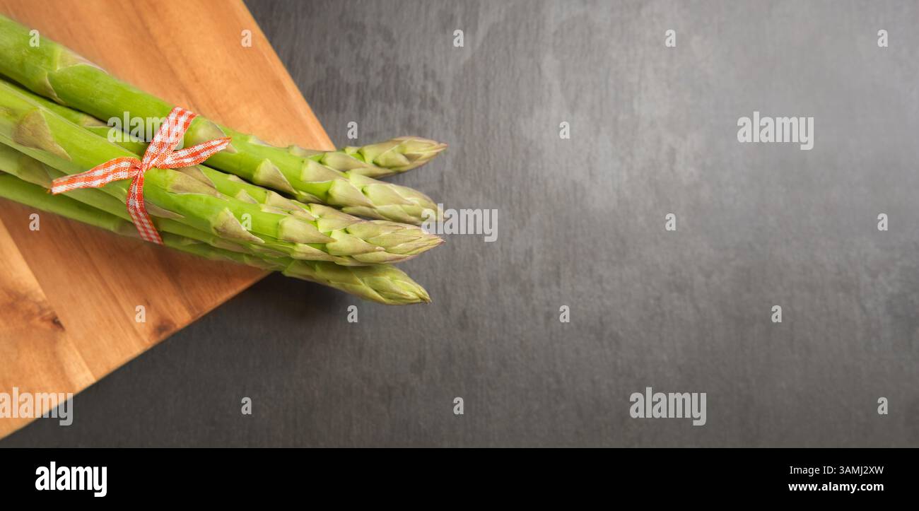Les lances d'asperges vertes se rapprochent. Un bouquet d'asperges vertes crues fraîches sur une planche à découper en bois sur un fond gris avec bannière de vue de dessus d'espace de copie. Banque D'Images