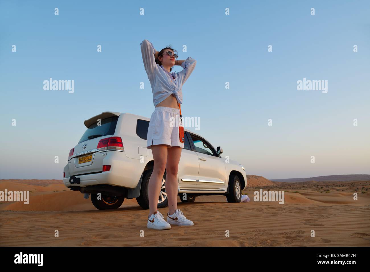 Rub Al Khali désert, Sultanat d'Oman - 5 avril 2025 : jeune fille attrayante reste près de 4x4 de voiture de route dans le désert de Wahiba Sands au lever du soleil, armes u Banque D'Images