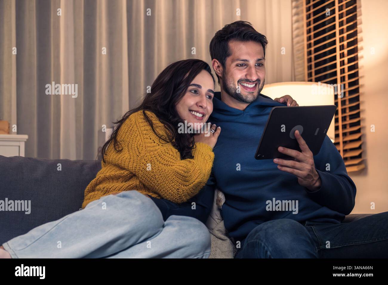 Couple hispanique utilisant une tablette numérique pour diffuser un spectacle ensemble à la maison sur le canapé la nuit Banque D'Images