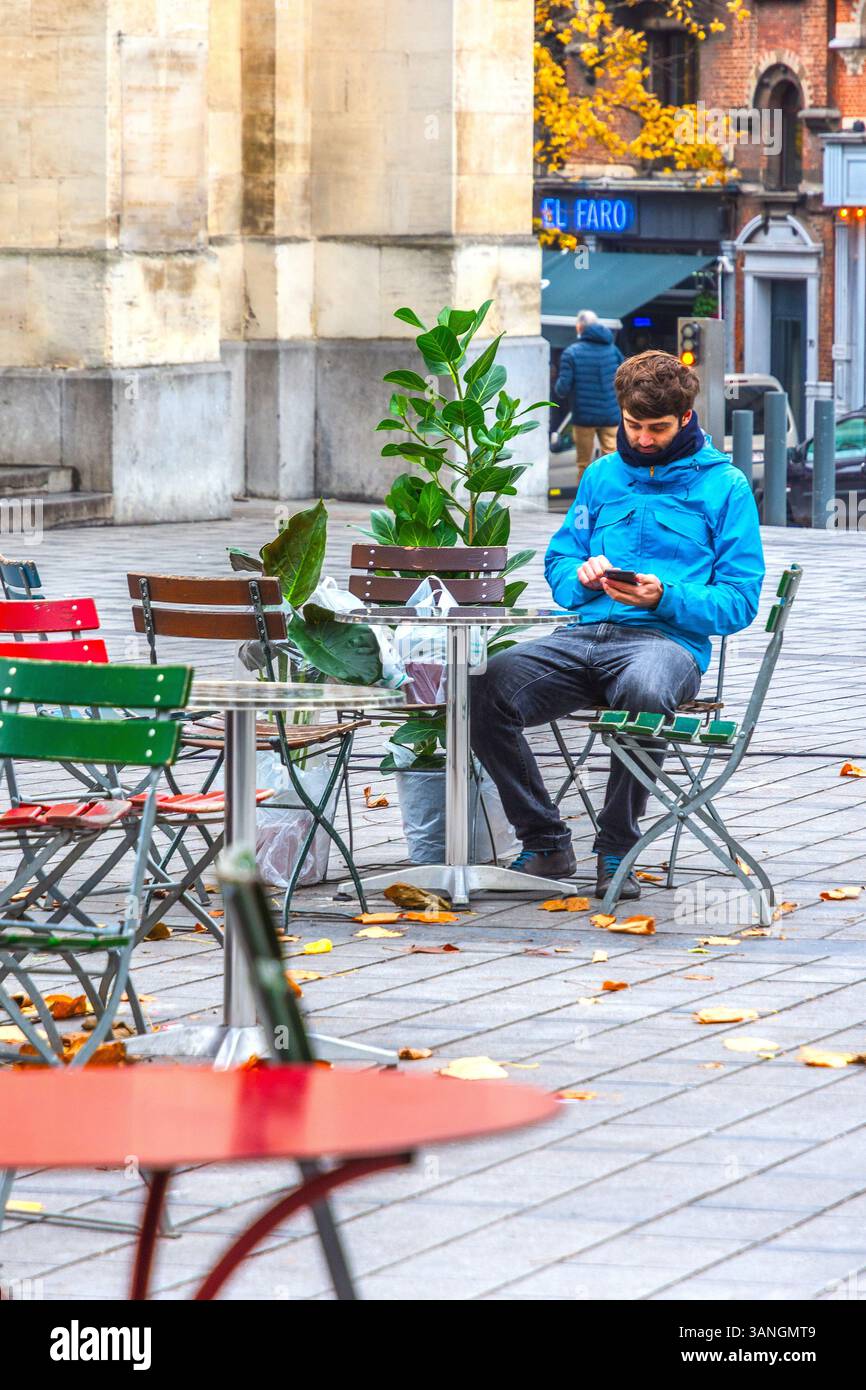 Homme avec deux grandes plantes en pot assis à la table de café en plein air vérifiant smartphone - Bruxelles, Belgique. Banque D'Images