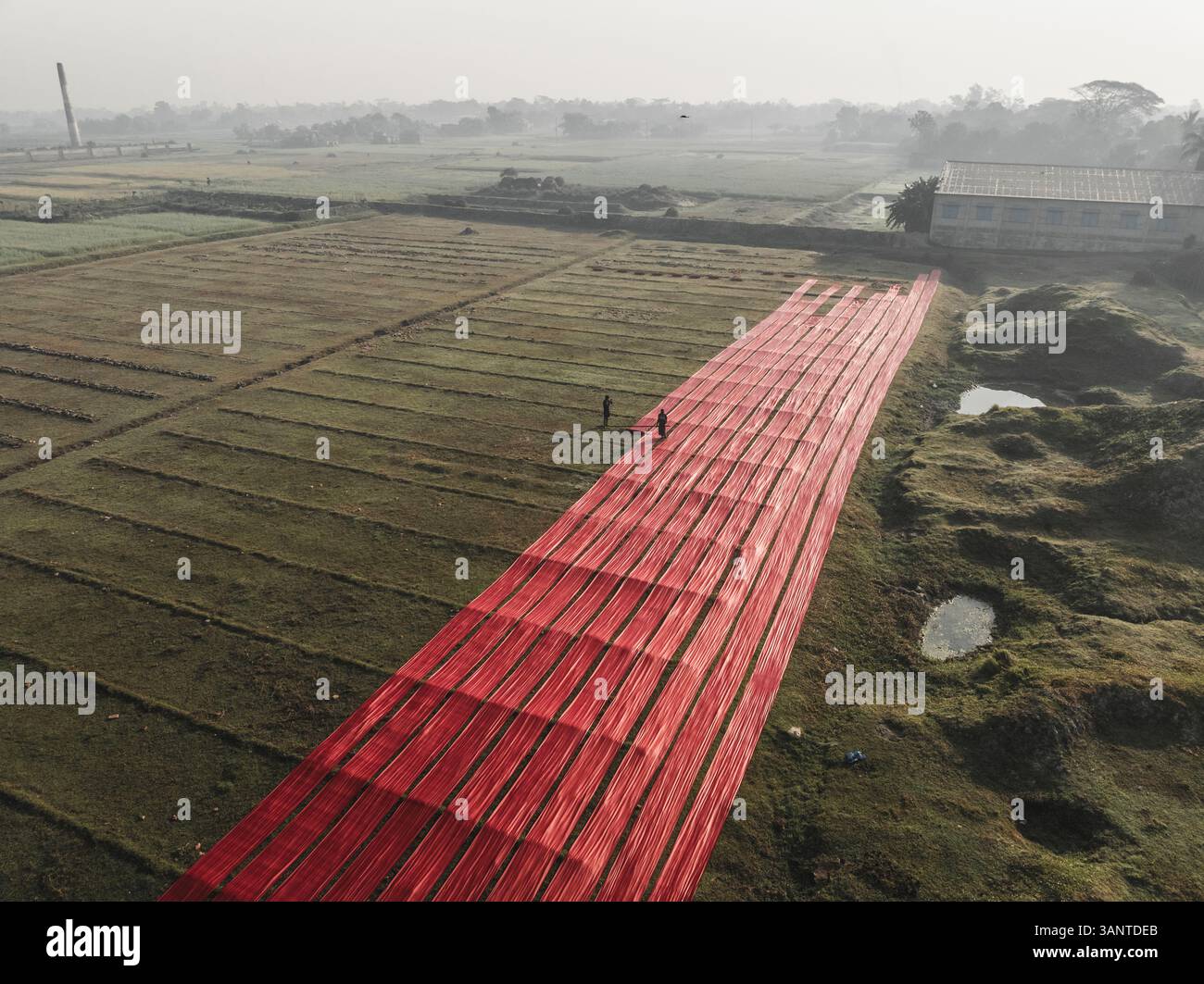 Vue aérienne de personnes séchant des tissus dans une usine de tissus entourée de champs ruraux, Paikar Char, Narsingdi, Bangladesh. Banque D'Images