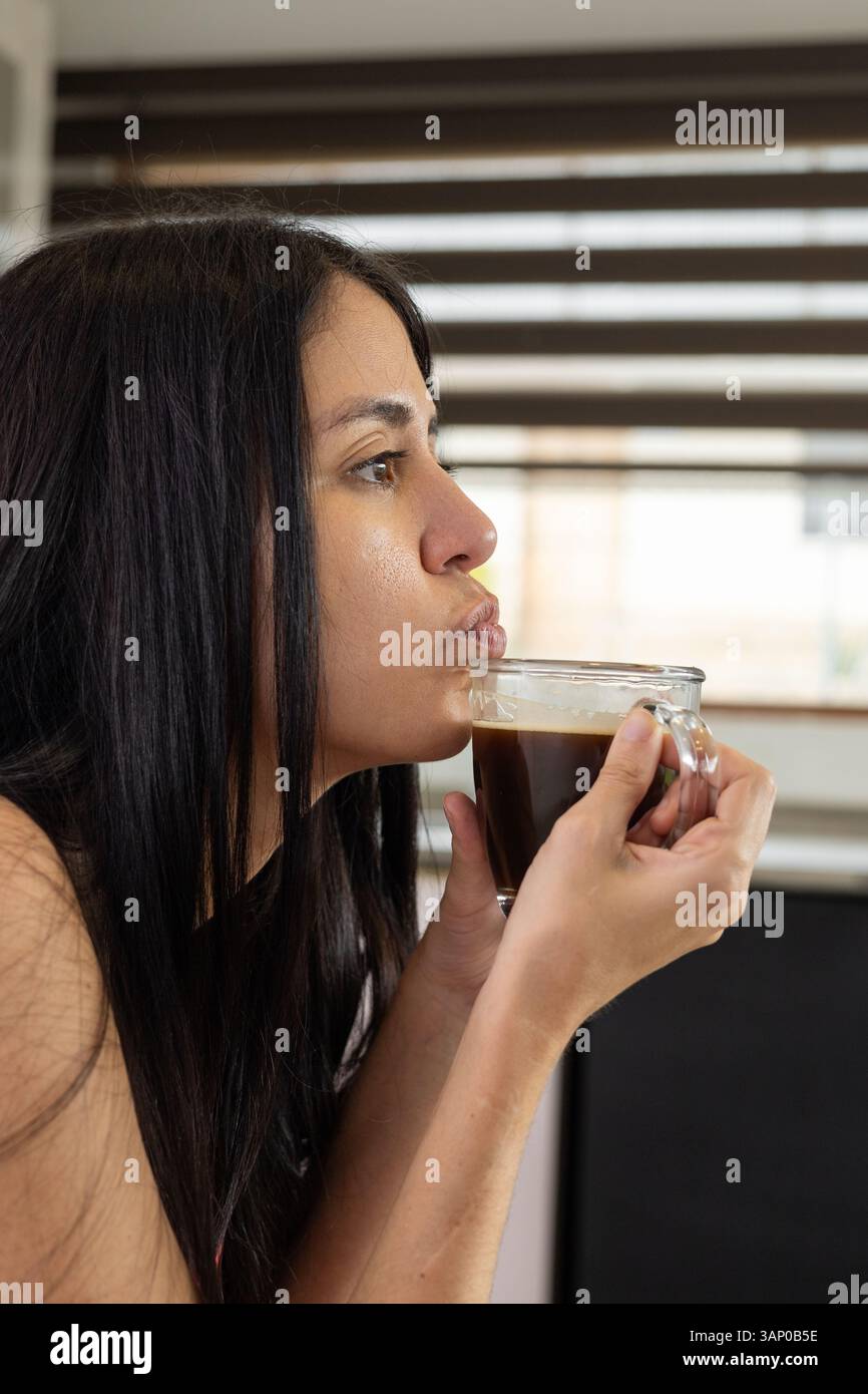 Une jeune femme souffle doucement sur sa tasse à café, attendant qu'elle refroidisse, alors qu'elle profite d'un moment calme et réfléchi dans un cadre confortable. Banque D'Images