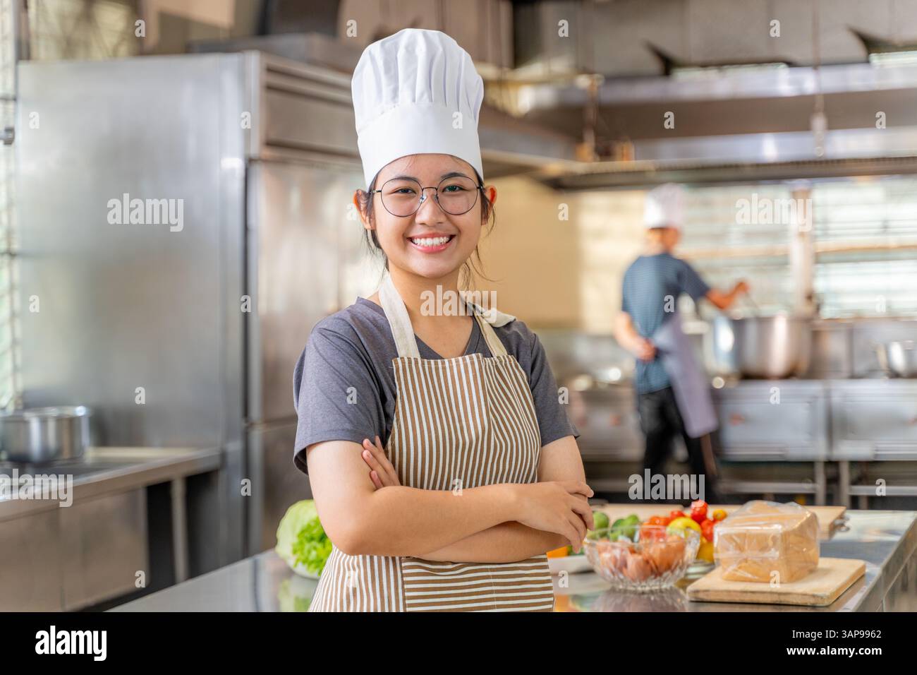 Jeune femme chef confiante souriante et debout avec les bras croisés dans une cuisine commerciale. Elle porte un chapeau de chef traditionnel et un tablier rayé, avec f Banque D'Images