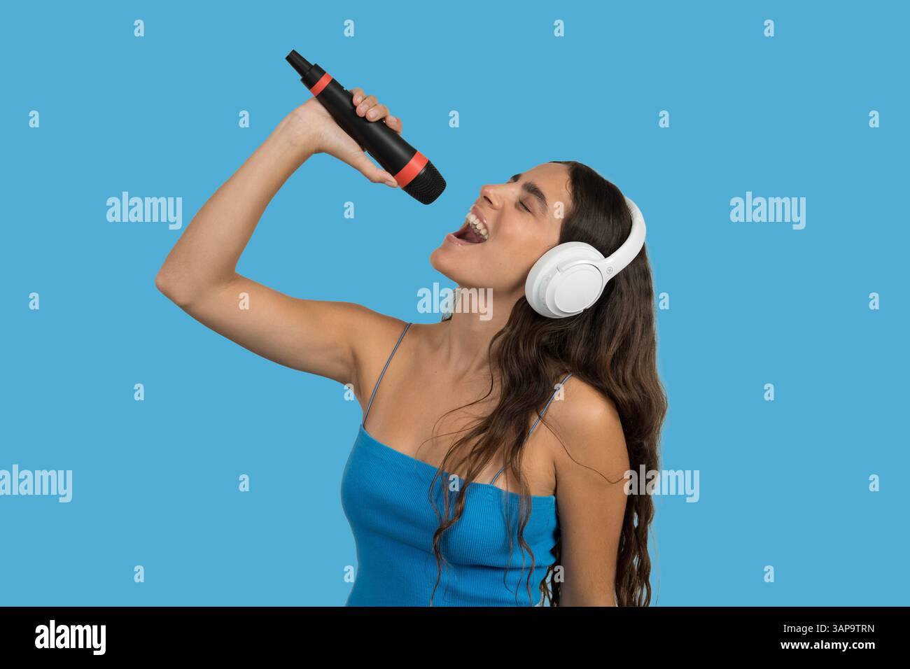 Chanteuse hispanique passionnée appréciant la musique, jouant avec un casque sans fil et un microphone en studio avec fond de couleur unie Banque D'Images