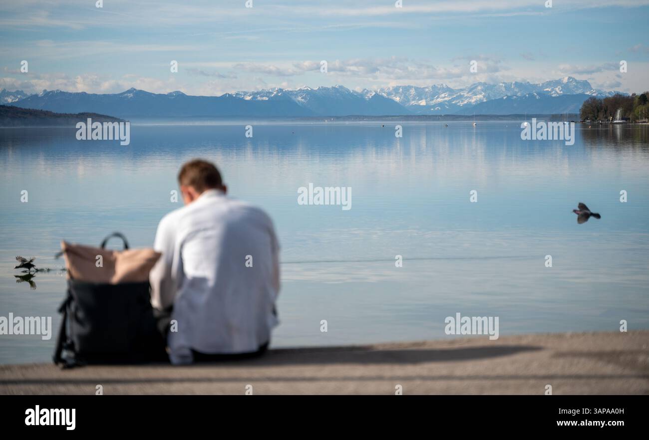 Starnberg, Allemagne. 16 avril 2025. Un homme est assis sur les rives du lac Starnberg devant un panorama montagneux des Alpes bavaroises enneigées. Crédit : Leonie Asendorpf/dpa/Alamy Live News Banque D'Images