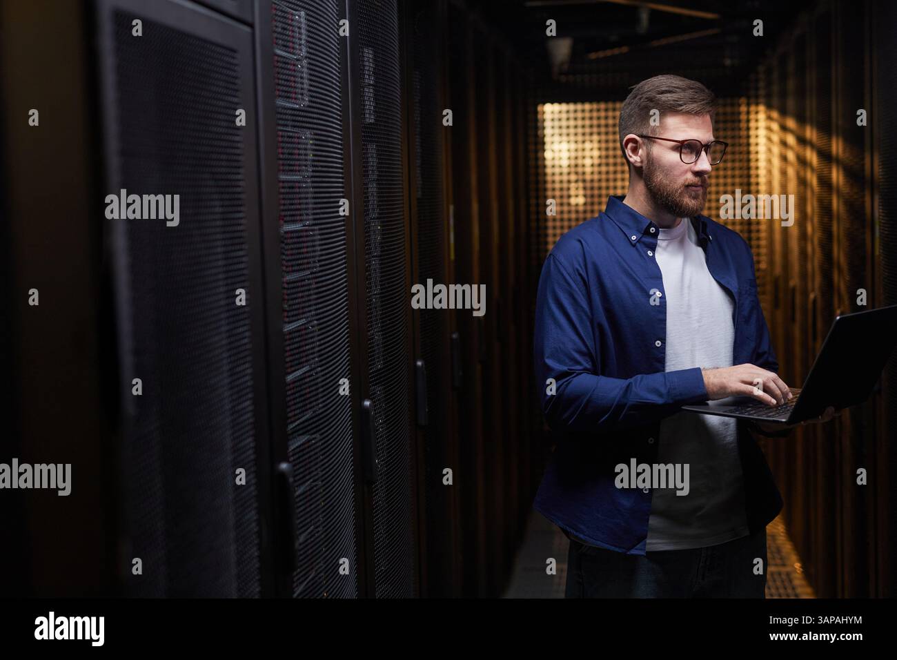 Homme exploitant un ordinateur portable dans un centre de données entouré de racks de serveurs et d'équipements. Surveillance professionnelle concentrée des systèmes de réseau et garantie d'un bon fonctionnement Banque D'Images