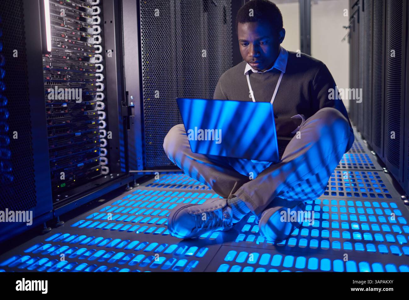 Homme assis au sol dans le centre de données, exploitant un ordinateur portable au milieu des racks de serveurs. Des lumières LED bleues éclairent la zone environnante, créant une ambiance futuriste Banque D'Images