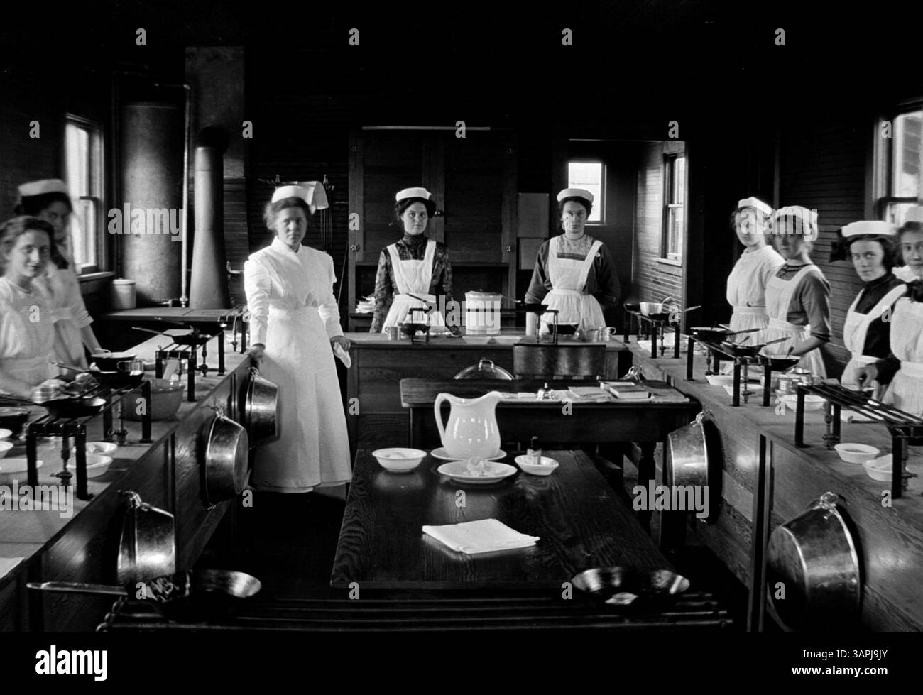 Cette image en noir et blanc montre un groupe de femmes portant des tabliers et des chapeaux blancs, probablement des étudiantes en économie domestique à Eugene, Oregon, debout à côté de comptoirs remplis d'appareils de chauffage et de casseroles. Banque D'Images