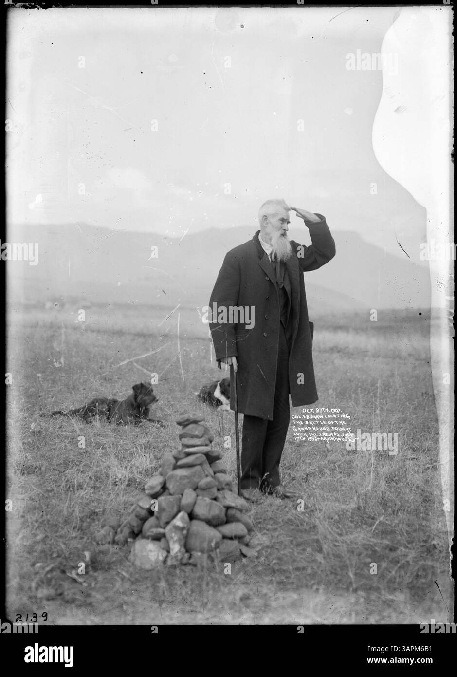 Photographie prise le 27 octobre 1907 du colonel F.B. Shaw localisant le site de la bataille du Grand Round, combattu avec les Amérindiens le 17 juillet 1856. Shaw, un homme âgé en costume de laine, est vu dans un champ herbeux avec des chiens et un cairn de roche, avec une chaîne de montagnes en arrière-plan. Banque D'Images