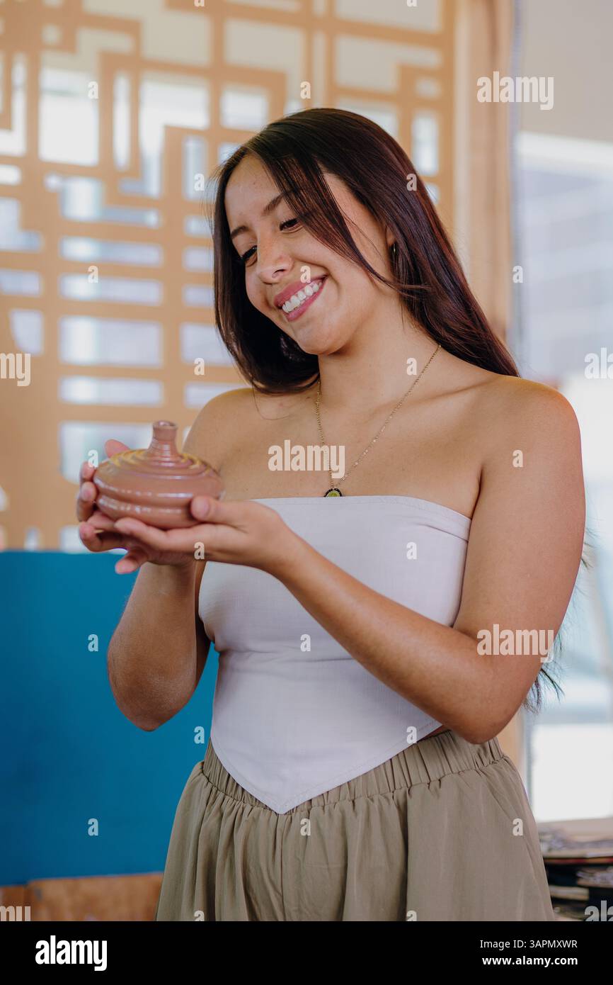 La jeune femme latina tient et évalue les pièces en céramique qu'elle a fabriquées dans son atelier. Entourée d’outils, elle sourit chaleureusement et porte des vêtements semi-formels. Banque D'Images