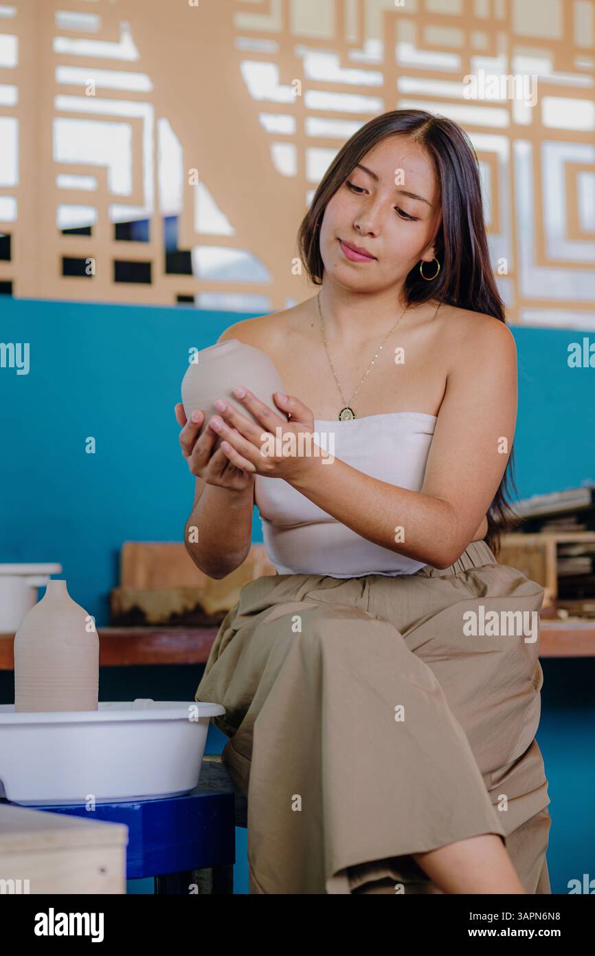 La jeune femme latina tient et évalue les pièces en céramique qu'elle a fabriquées dans son atelier. Entourée d’outils, elle sourit chaleureusement et porte des vêtements semi-formels. Banque D'Images