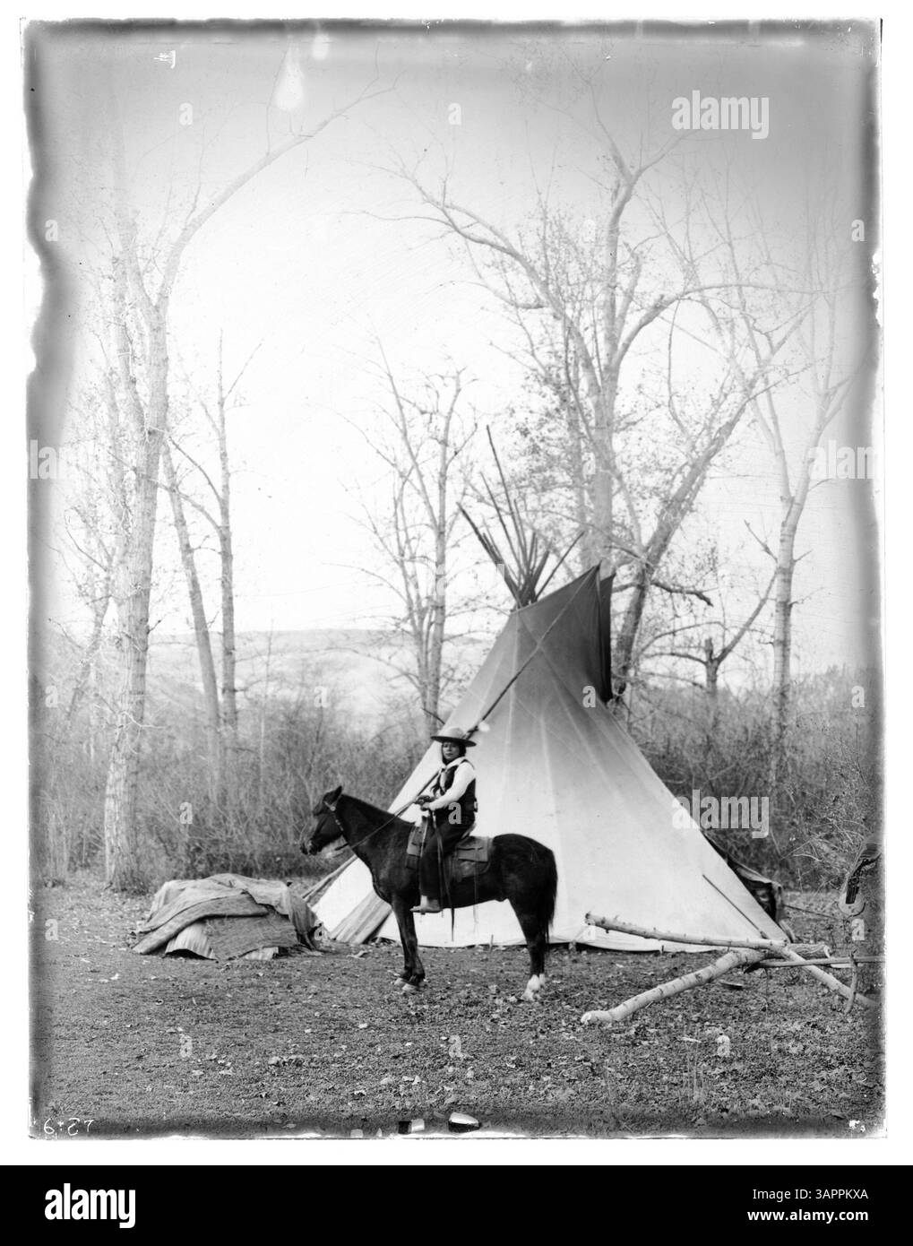 Un amérindien portant des vêtements euro-américains est assis sur un cheval, avec un tipi et des couvertures en arrière-plan. La photographie est placée dans un paysage naturel avec des arbres, des arbustes et des montagnes lointaines. Banque D'Images