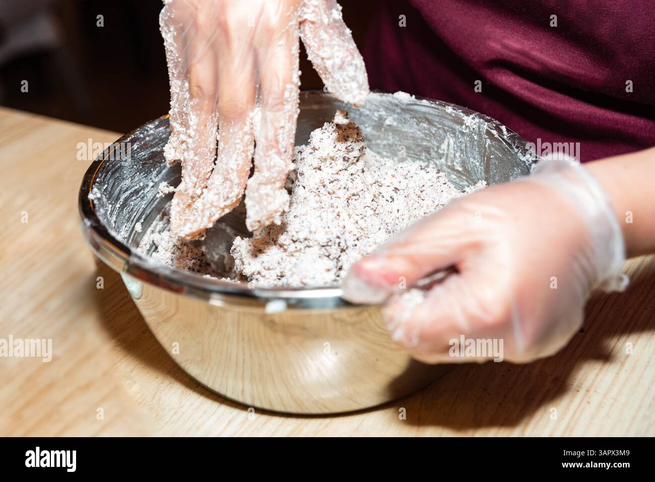 Gros plan d'une personne portant des gants tout en mélangeant la farine et le mélange de cacao dans la pâte à l'intérieur d'un bol de mélange en acier inoxydable. Banque D'Images