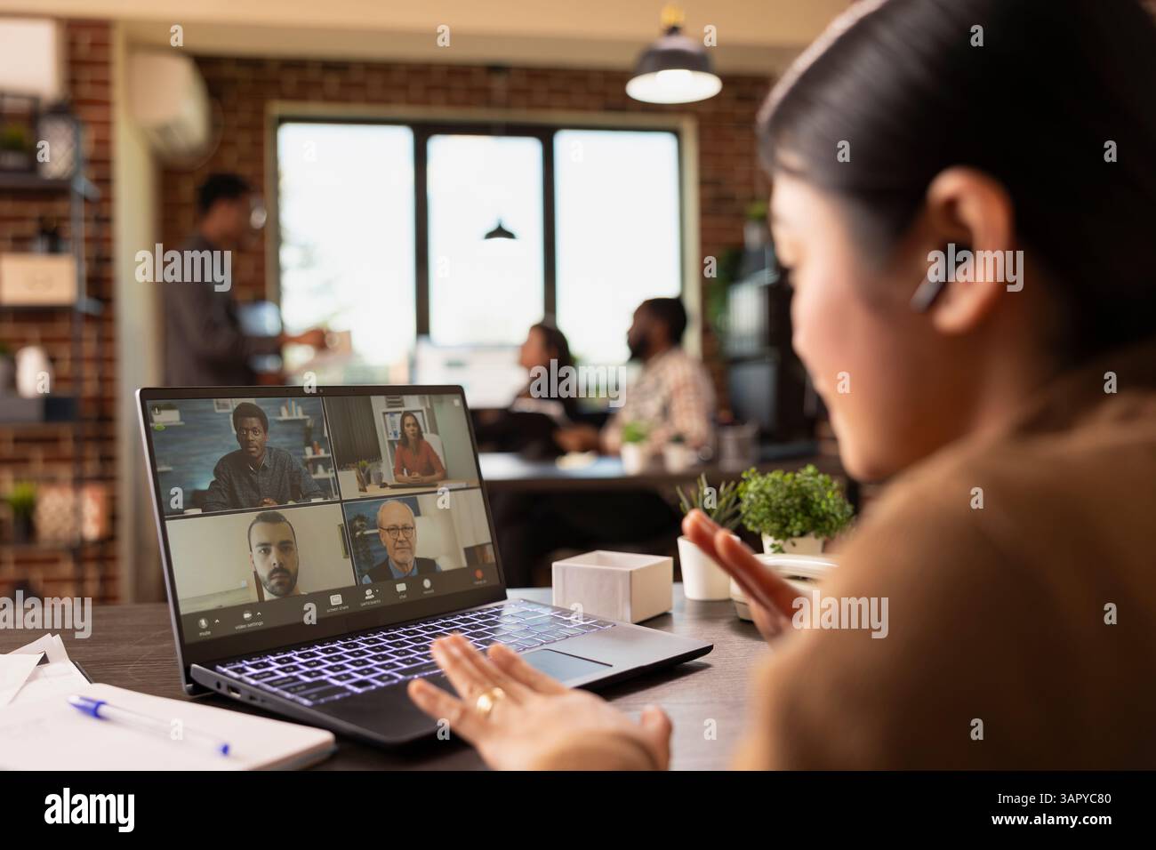 Gros plan d'un ordinateur portable affichant une vidéoconférence avec des collègues, tandis qu'une femme d'affaires avec des écouteurs s'engage dans la discussion. La manager asiatique assiste à une réunion en ligne depuis son bureau. Banque D'Images