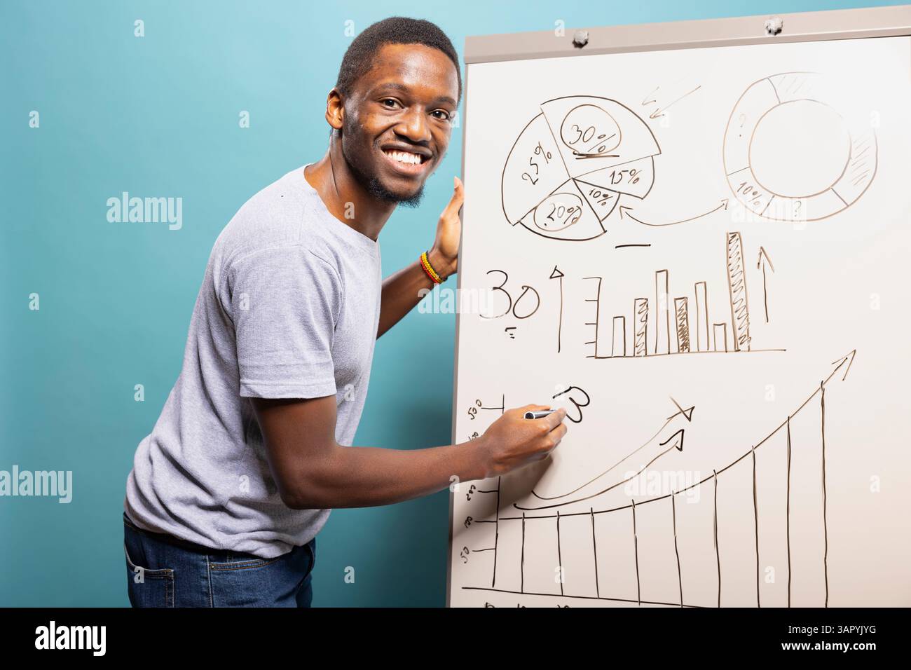Jeune homme noir écrivant sur un tableau blanc et expliquant un graphique, debout sur un fond bleu de studio. Souriant afro-américain tenant un stylo et utilisant un tableau de marqueurs pour présenter un graphique. Banque D'Images