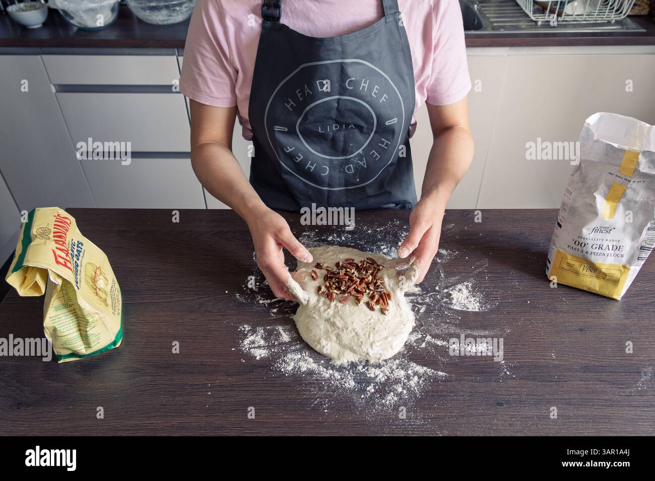 Une femme dans la trentaine pétrissant de la pâte, portant un tablier et faisant du pain maison avec des noix de pécan, Royaume-Uni. Concept : cuisson maison, nourriture de confort, style de vie Banque D'Images