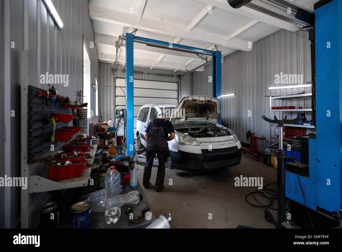 Un mécanicien travaille sur une fourgonnette commerciale blanche avec son capot ouvert à l'intérieur d'un atelier de réparation automobile occupé équipé d'un élévateur de véhicule et de divers outils Banque D'Images