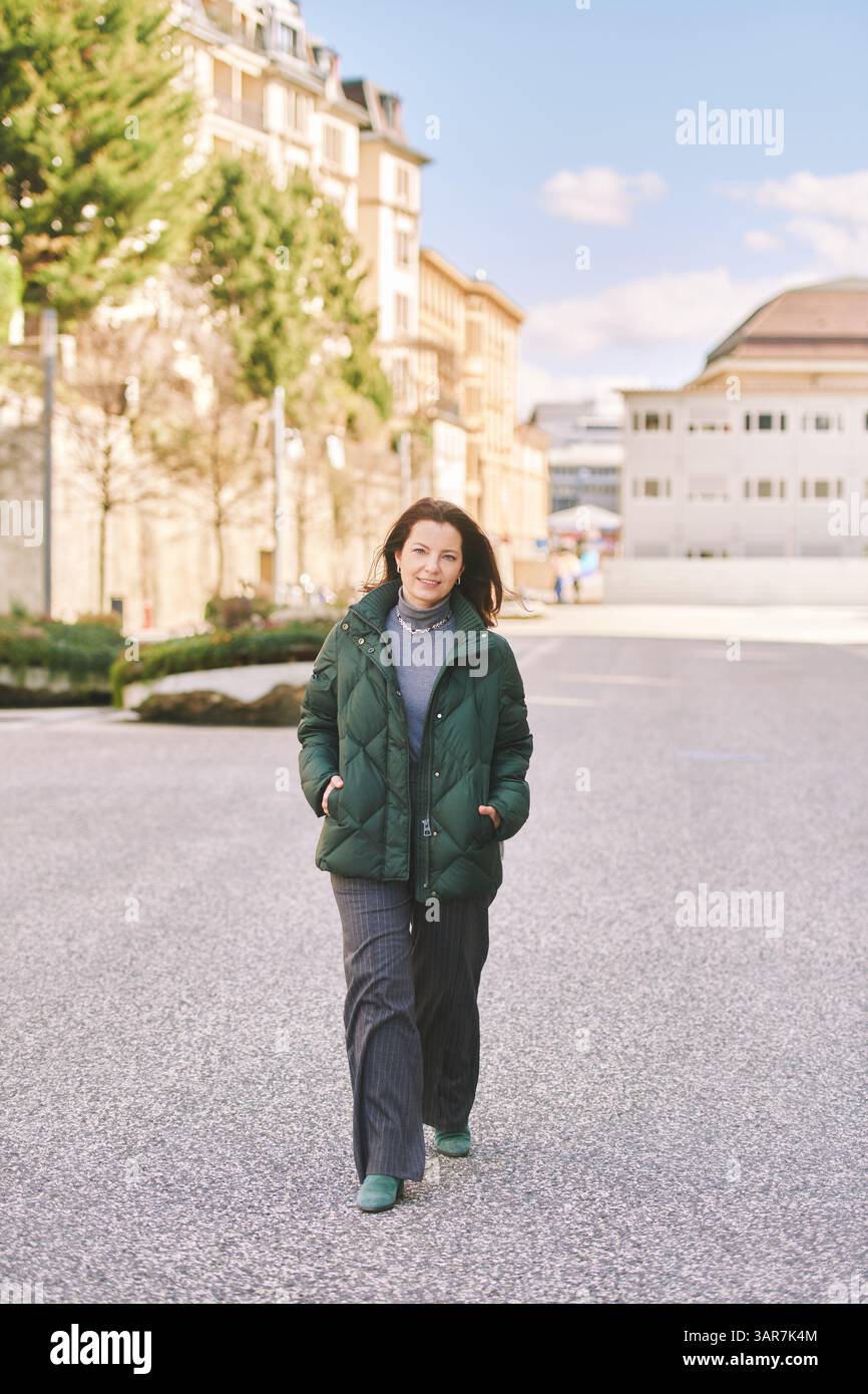 Plein air plein - portrait en longueur d'une femme heureuse d'âge moyen marchant dans la rue, portant une veste verte chaude Banque D'Images