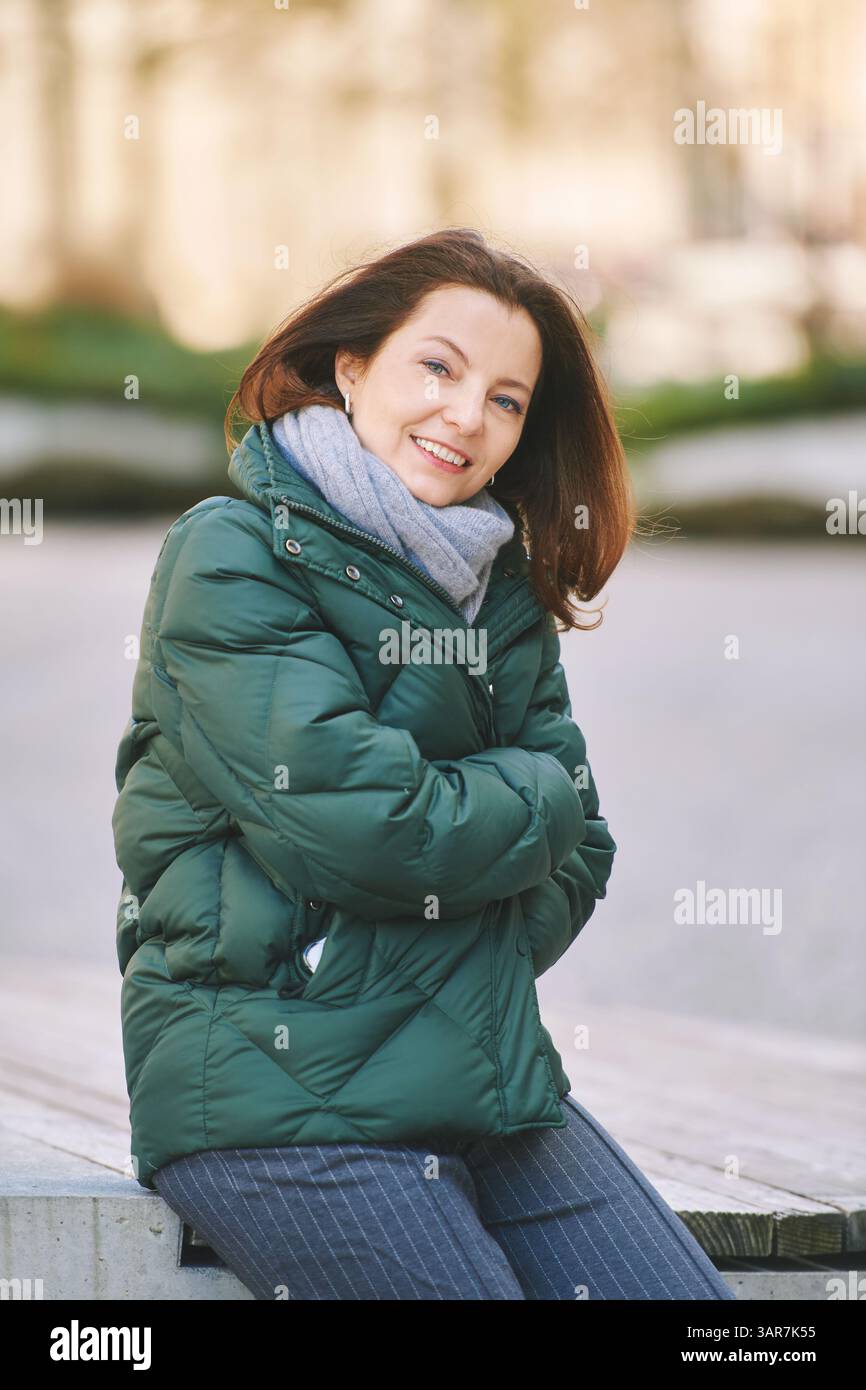 Portrait extérieur d'une femme heureuse assise sur le banc, portant une veste verte chaude Banque D'Images