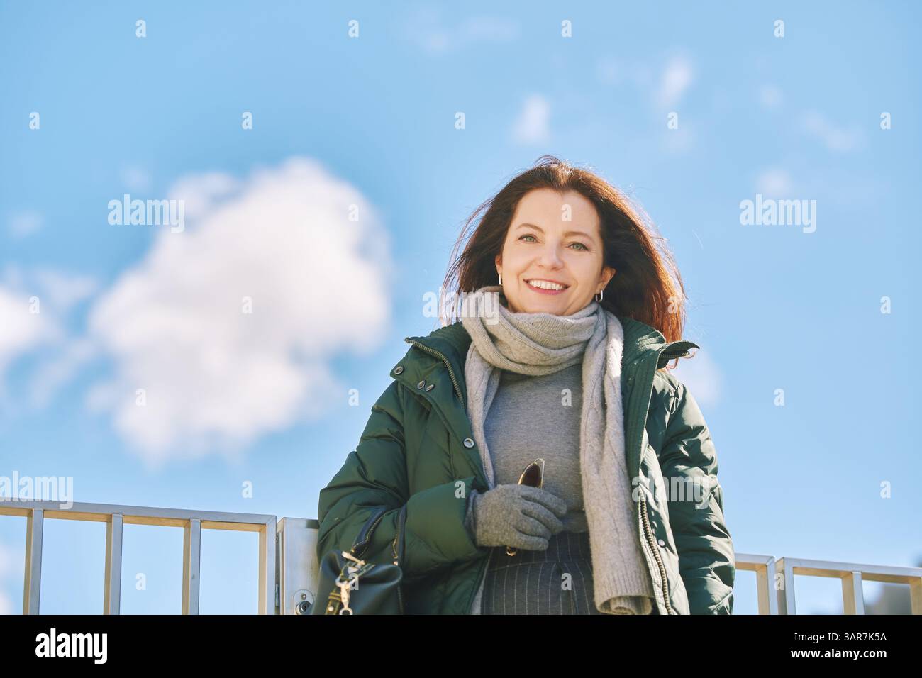 Portrait extérieur de belle femme d'âge moyen posant contre le ciel bleu, portant une veste verte chaude et une écharpe grise avec des gants, temps frais Banque D'Images