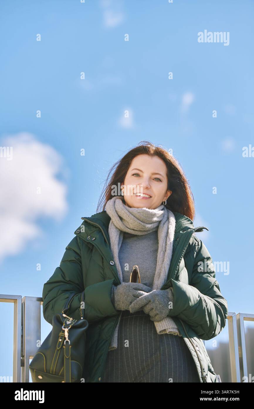 Portrait extérieur de belle femme d'âge moyen posant contre le ciel bleu, portant une veste verte chaude et une écharpe grise avec des gants, temps frais Banque D'Images