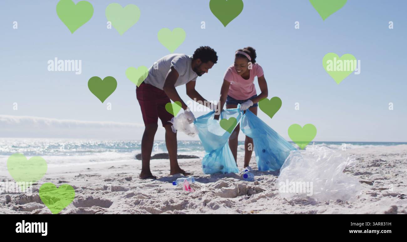 Image de coeurs verts sur un couple afro-américain avec des sacs de recyclage nettoyant la plage Banque D'Images