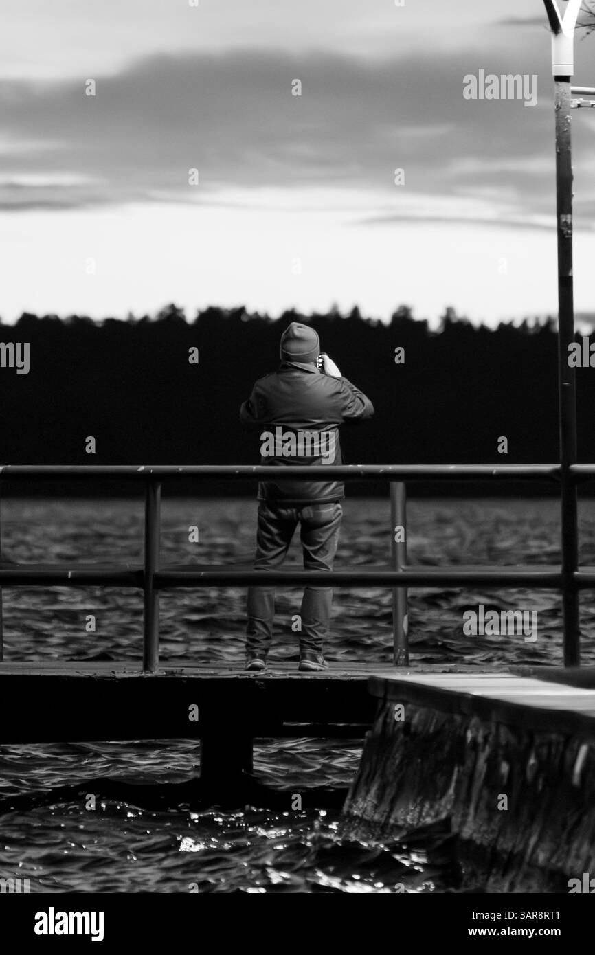 Homme anonyme près d'un lac prenant des photos avec son téléphone portable du coucher du soleil. Photo noir et blanc en soirée. La personne est éclairée. Banque D'Images