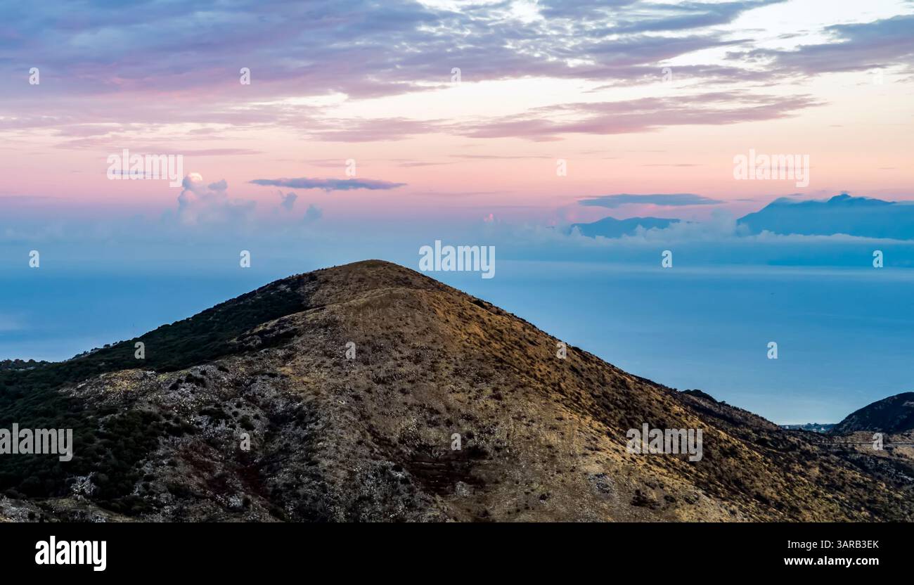 Un paysage serein avec une colline en pente douce contre un ciel pastel au coucher du soleil. L'horizon rencontre la mer calme, avec des montagnes lointaines visibles. Banque D'Images