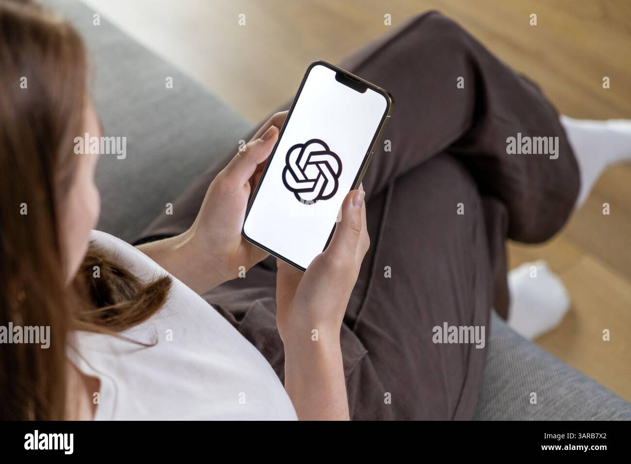 Poznan, Pologne – 14 avril 2025 : utilisatrice féminine se relaxant sur un canapé à la maison tout en tenant un smartphone avec le logo ChatGPT, représentant une utilisation occasionnelle du châssis ai Banque D'Images