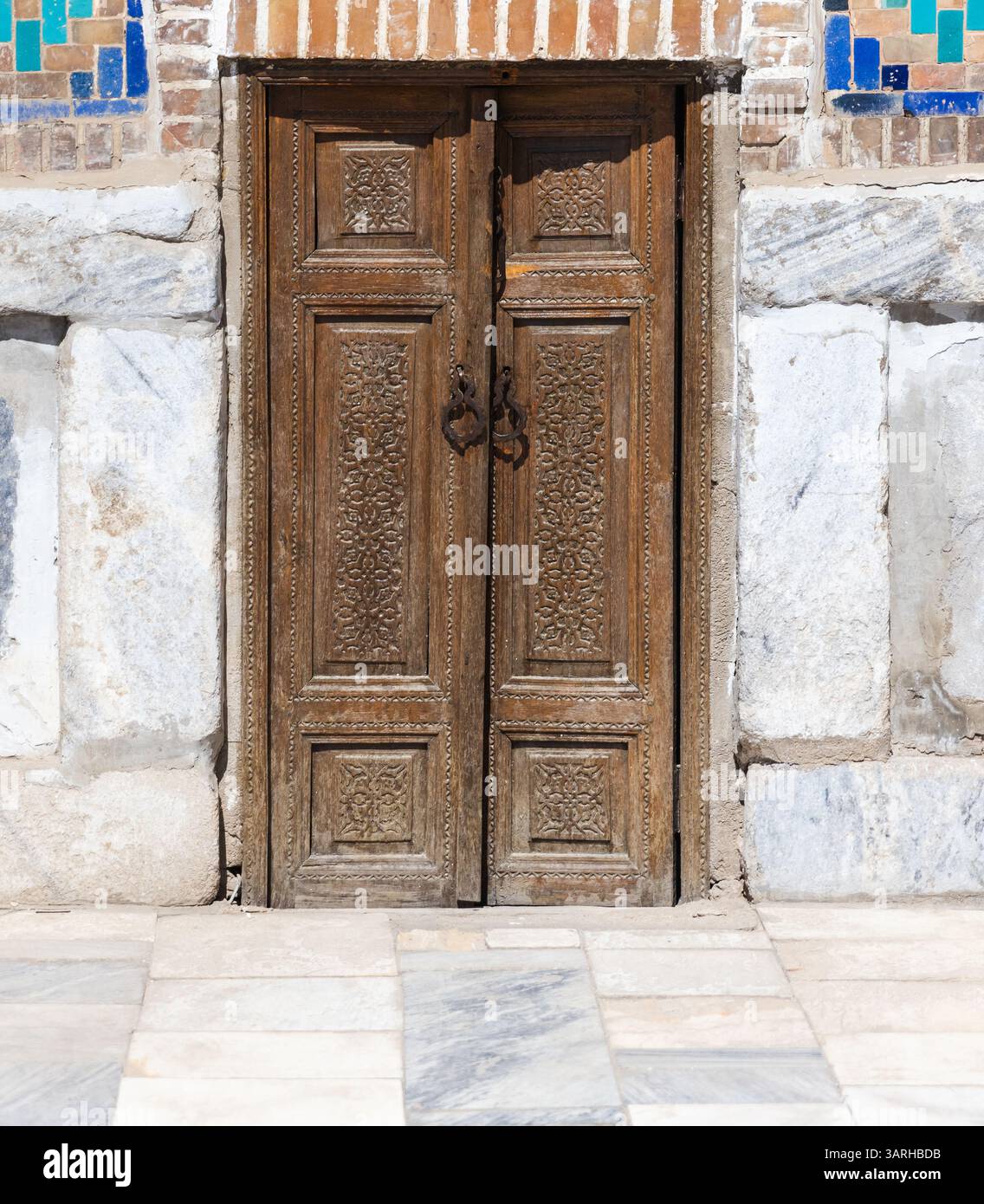 Des portes en bois au design complexe sont placées dans un mur de pierre et de briques avec un décor de tuiles vives, créant un mélange d'artisanat traditionnel et d'artisti Banque D'Images
