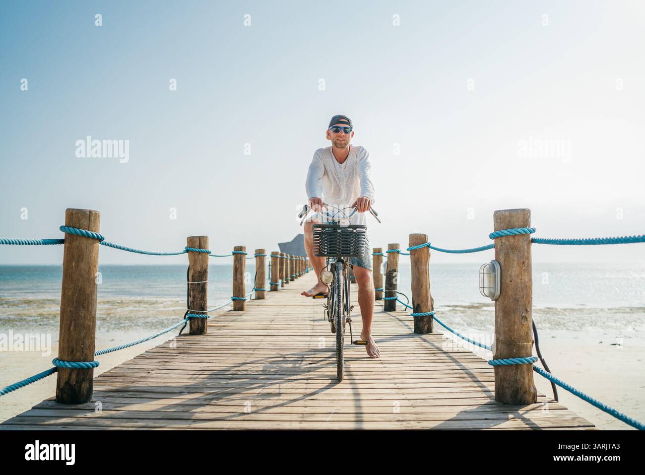 Portrait d'un homme heureux et souriant vêtu de vêtements d'été légers et de lunettes de soleil, à vélo sur une jetée en bois au bord de la mer. Des vacances sans soucis à Banque D'Images